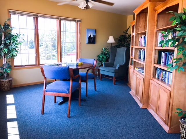 A cozy living room with wooden furniture, including a bookshelf and a table with chairs, illuminated by natural light from a large window.