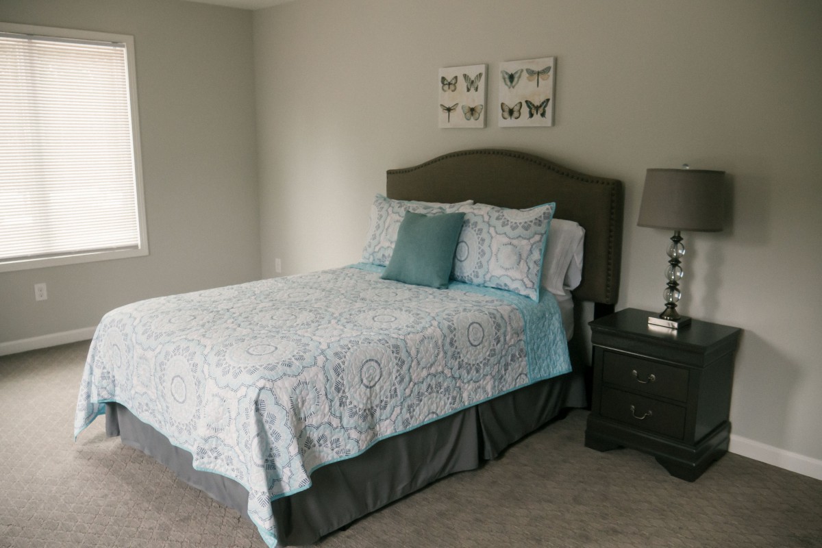 A neatly arranged bedroom featuring a bed with patterned bedding, pillows, a nightstand with a lamp, and wall decorations.
