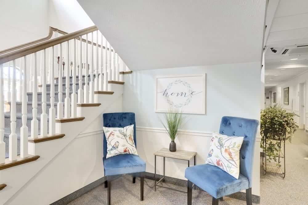 A cozy corner features two blue chairs with decorative pillows, a small side table with a plant, and wall art under a staircase.
