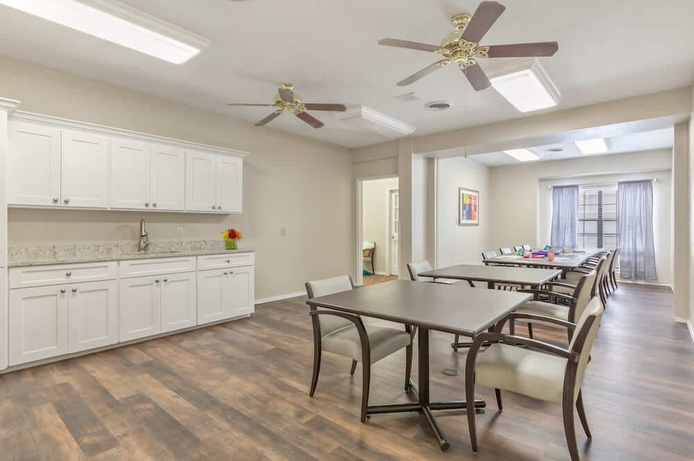 A spacious room with kitchen facilities, multiple tables, and ceiling fans.