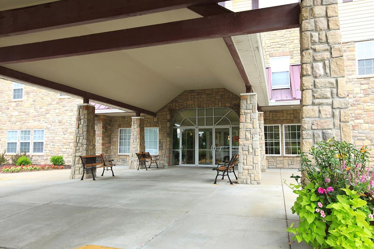 The image shows the entrance to a building with a covered walkway supported by stone pillars, accompanied by outdoor chairs.