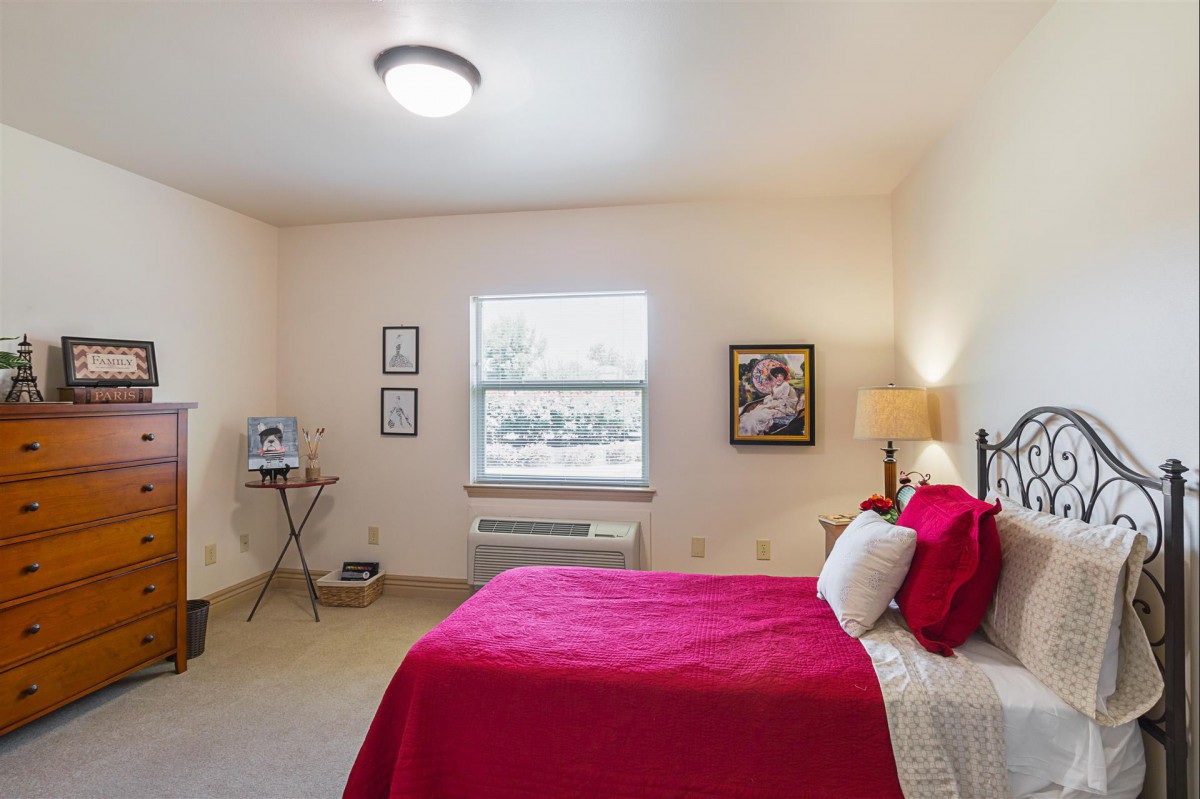 A cozy bedroom with a red bedspread, side table, and lamp.