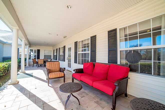 A covered patio with red and orange outdoor furniture, tables, and windows with shutters.