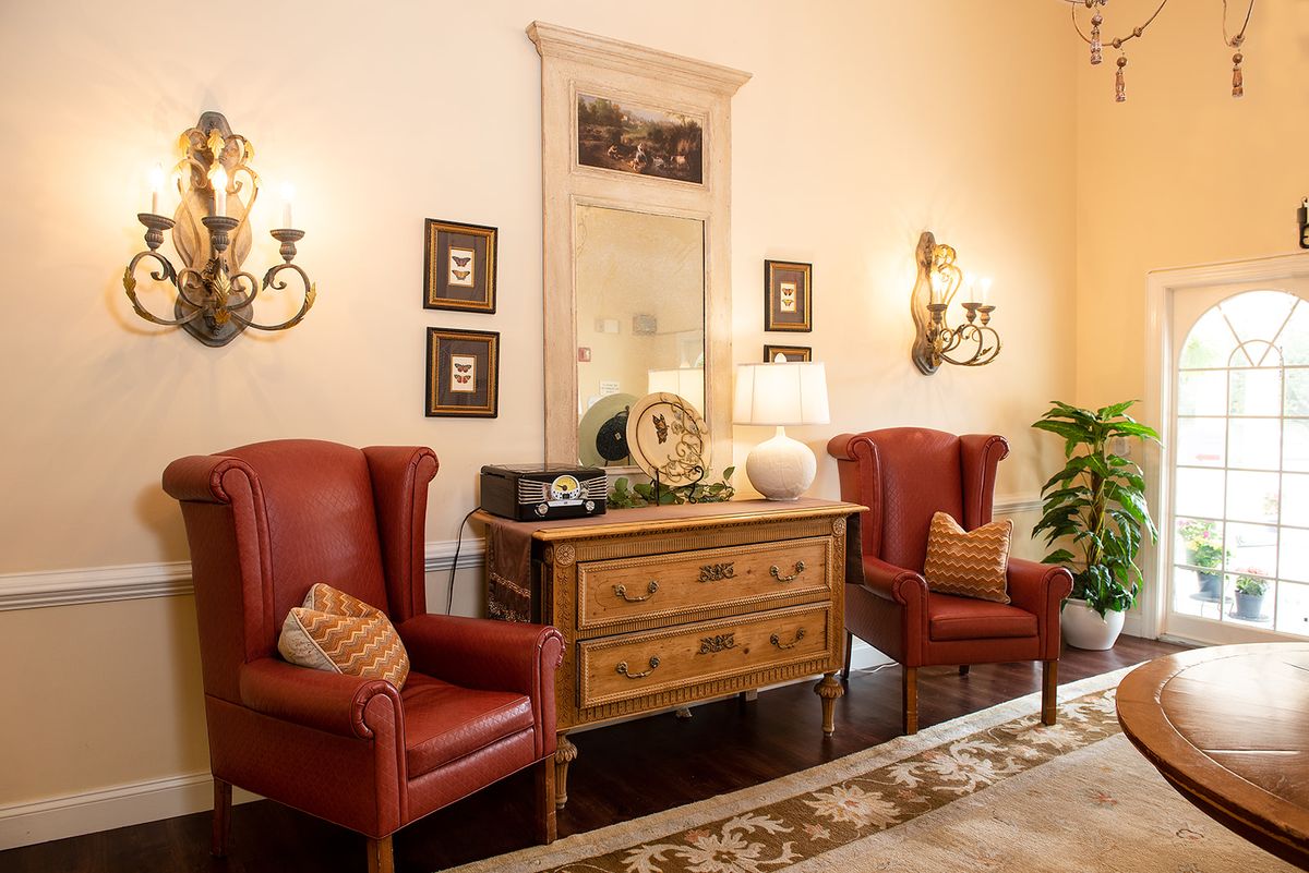 A cozy living room features vintage red armchairs and a wooden dresser with decorative items, enhanced by elegant wall sconces and a large mirror.