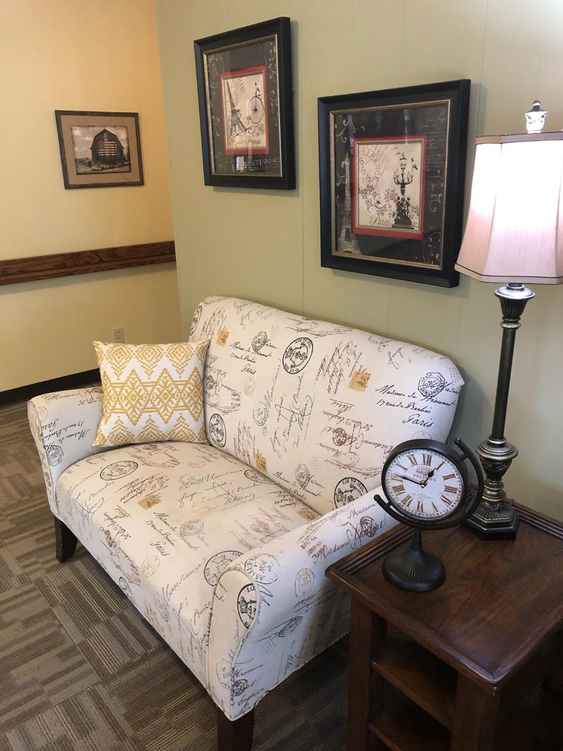 A cozy sitting area featuring a patterned sofa, decorative pillow, table lamp, and a vintage clock, complemented by wall art.