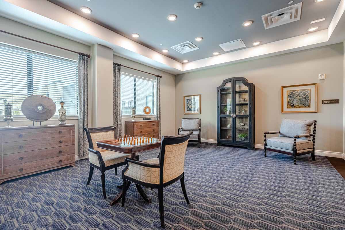 A cozy, well-lit room with a table set for chess, surrounded by comfortable chairs and a bookshelf.