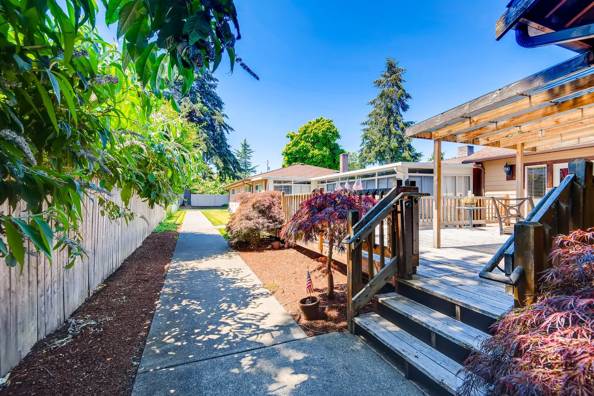 A sunny backyard featuring a wooden deck, a landscaped garden, and a pathway lined with trees and shrubs.