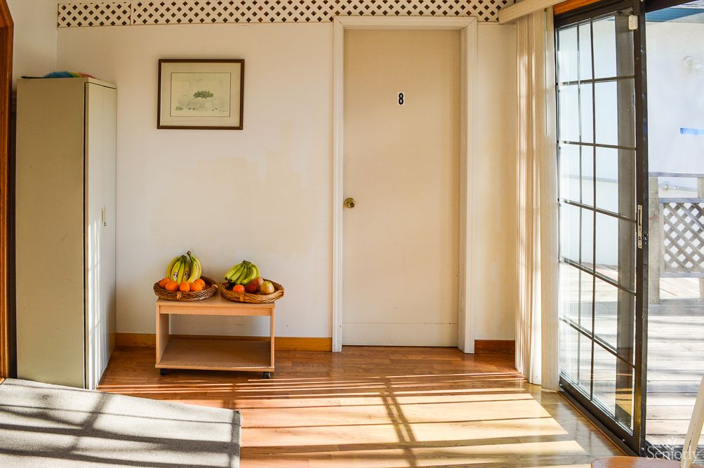 A sunlit room with a small table holding various fruits, a framed picture on the wall, and soft shadows cast across the floor.
