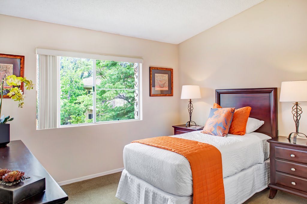 A cozy bedroom with a single bed and orange accents, featuring a large window that allows natural light to fill the space.