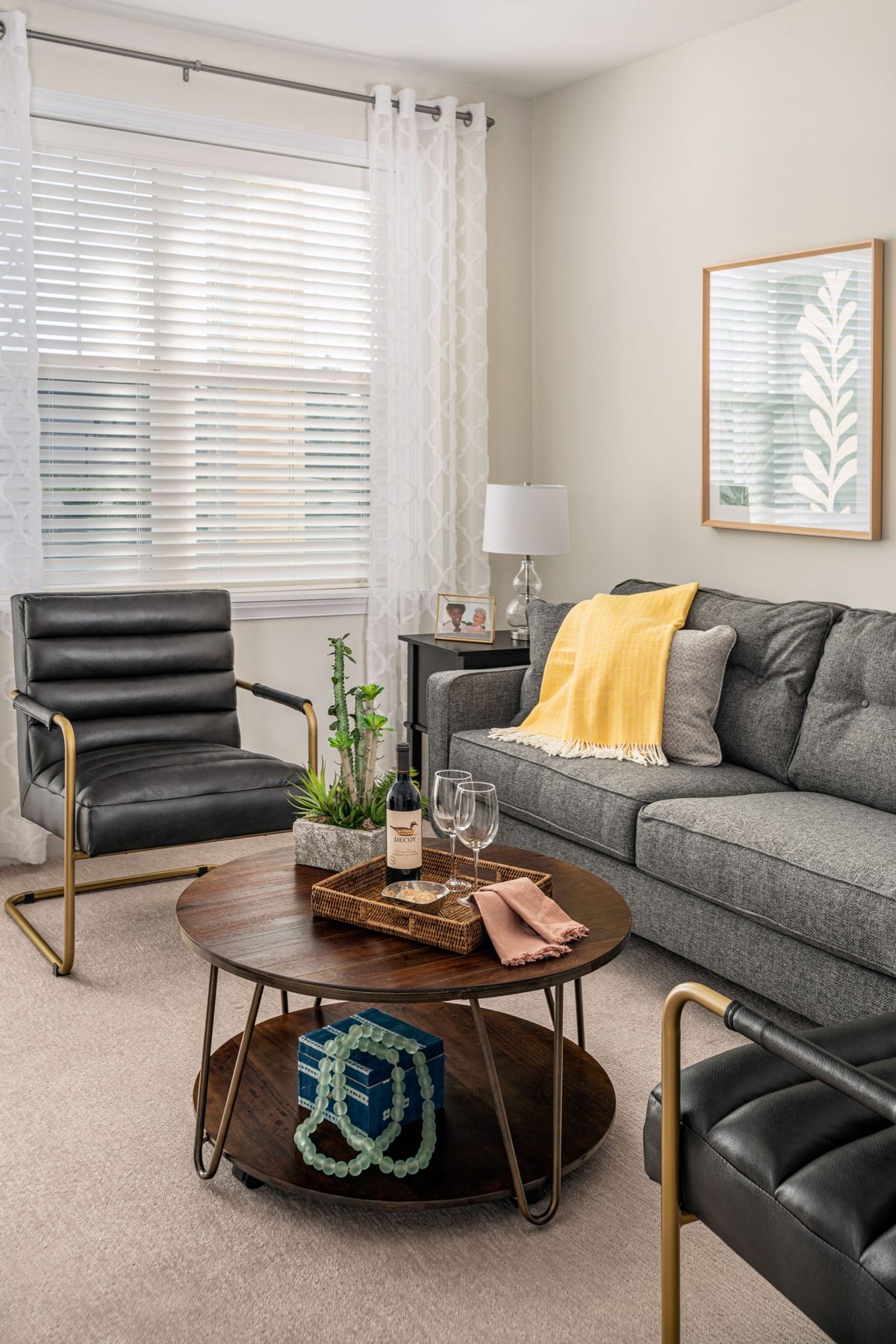 A cozy living room featuring a gray sofa with a yellow throw, a black armchair, and a round wooden coffee table adorned with decor items. The space is completed with wall art, a lamp, and green plants.