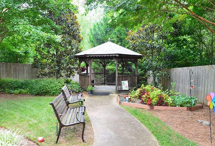 A tranquil garden setting featuring a gazebo, benches, and a pathway surrounded by lush greenery and colorful flowers.