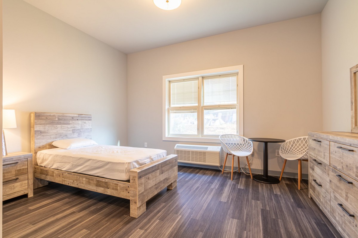 A minimalist bedroom with a single bed, window, table, and chairs on a hardwood floor.