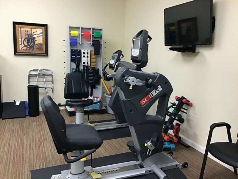 Image of a gym or rehabilitation facility featuring exercise equipment including a recumbent bike and weights.