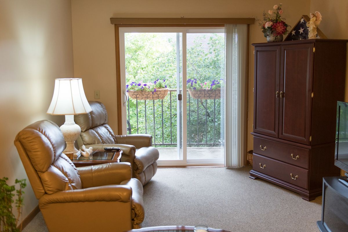 A cozy living room featuring armchairs, a lamp, and a sliding glass door leading to a small balcony with plants.