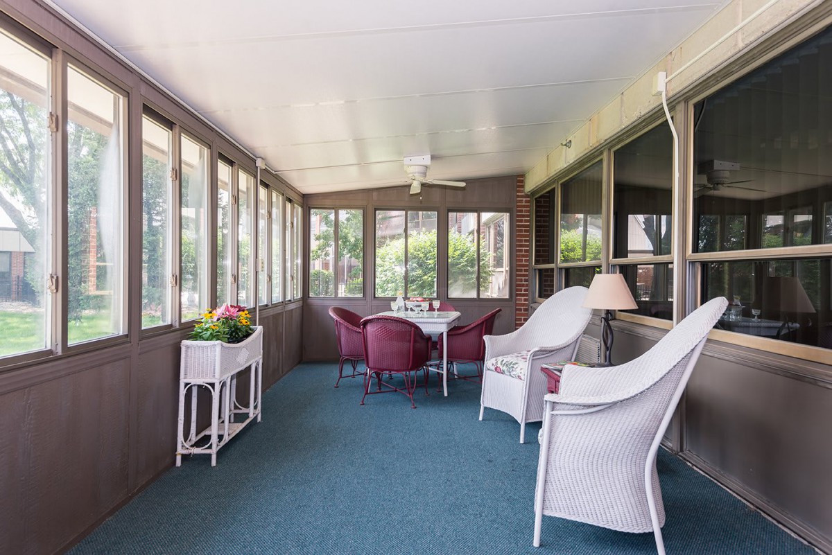 A cozy sunroom featuring wicker furniture and a small dining setup, surrounded by large windows and indoor plants.