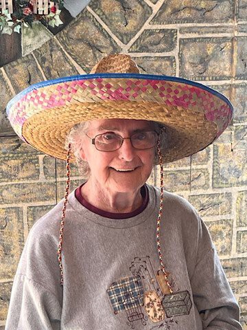 A woman wearing a sombrero and glasses, smiling indoors.
