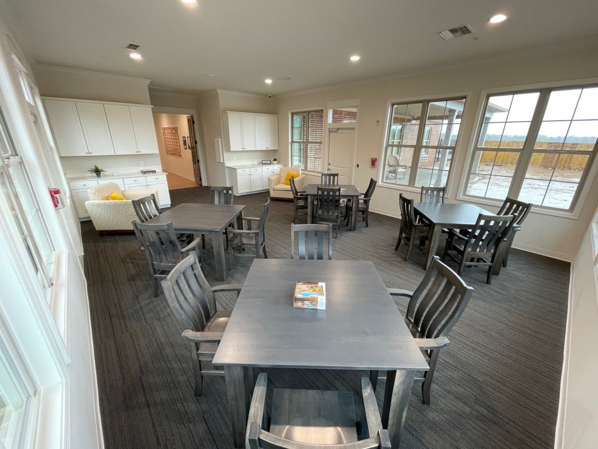 A modern communal dining area featuring multiple tables and chairs, a kitchenette, and large windows.