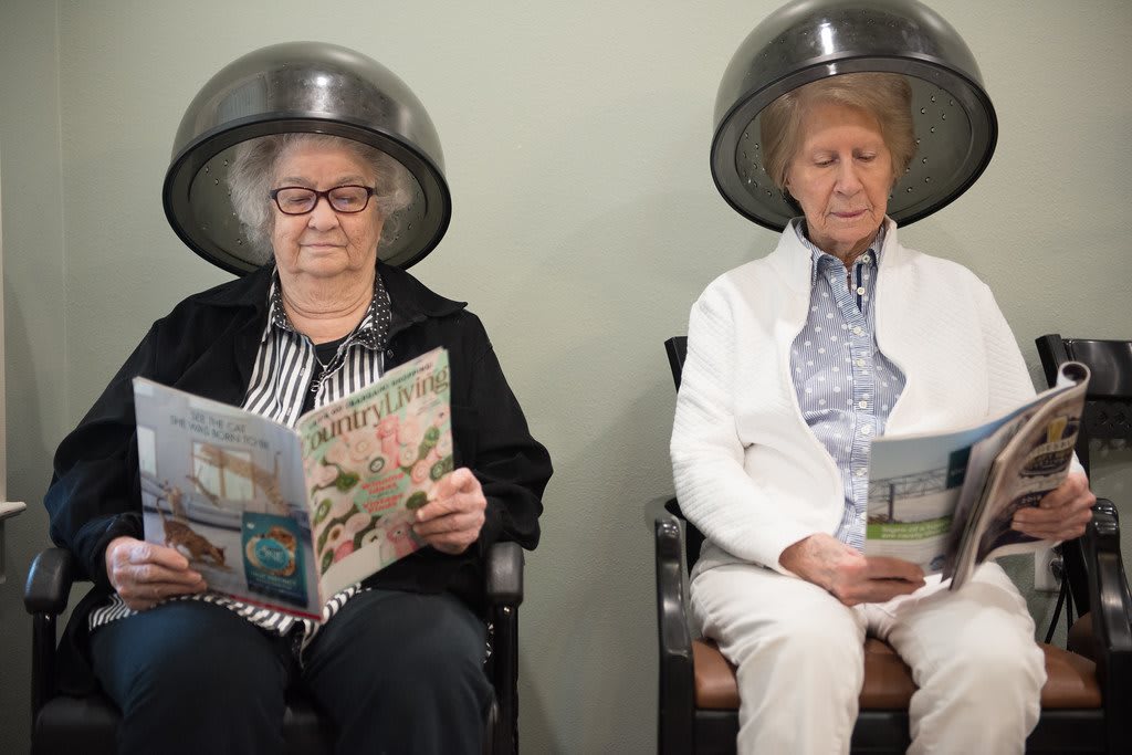 Two elderly women sitting under hair dryers in a salon, reading magazines.