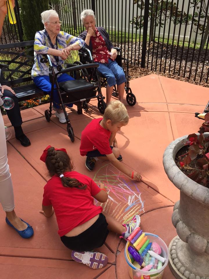 Children drawing with sidewalk chalk while adults sit nearby in a garden setting.