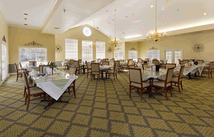 A spacious dining area with elegantly arranged tables and chairs, illuminated by chandeliers and natural light from large windows.