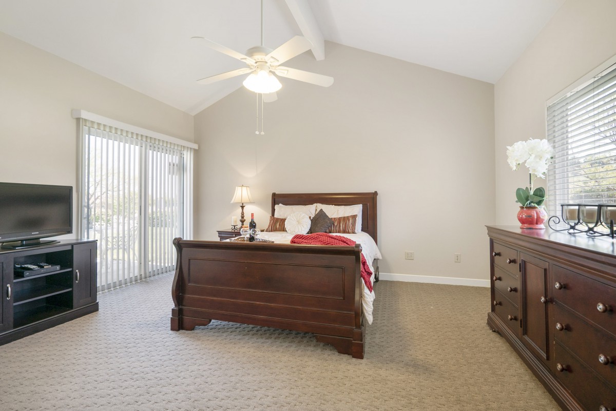 A cozy bedroom with a wooden bed frame, carpeted floor, and a ceiling fan.