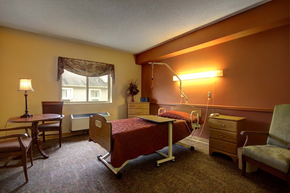 A well-lit hospital room featuring a bed and medical equipment, with furniture and a window view.