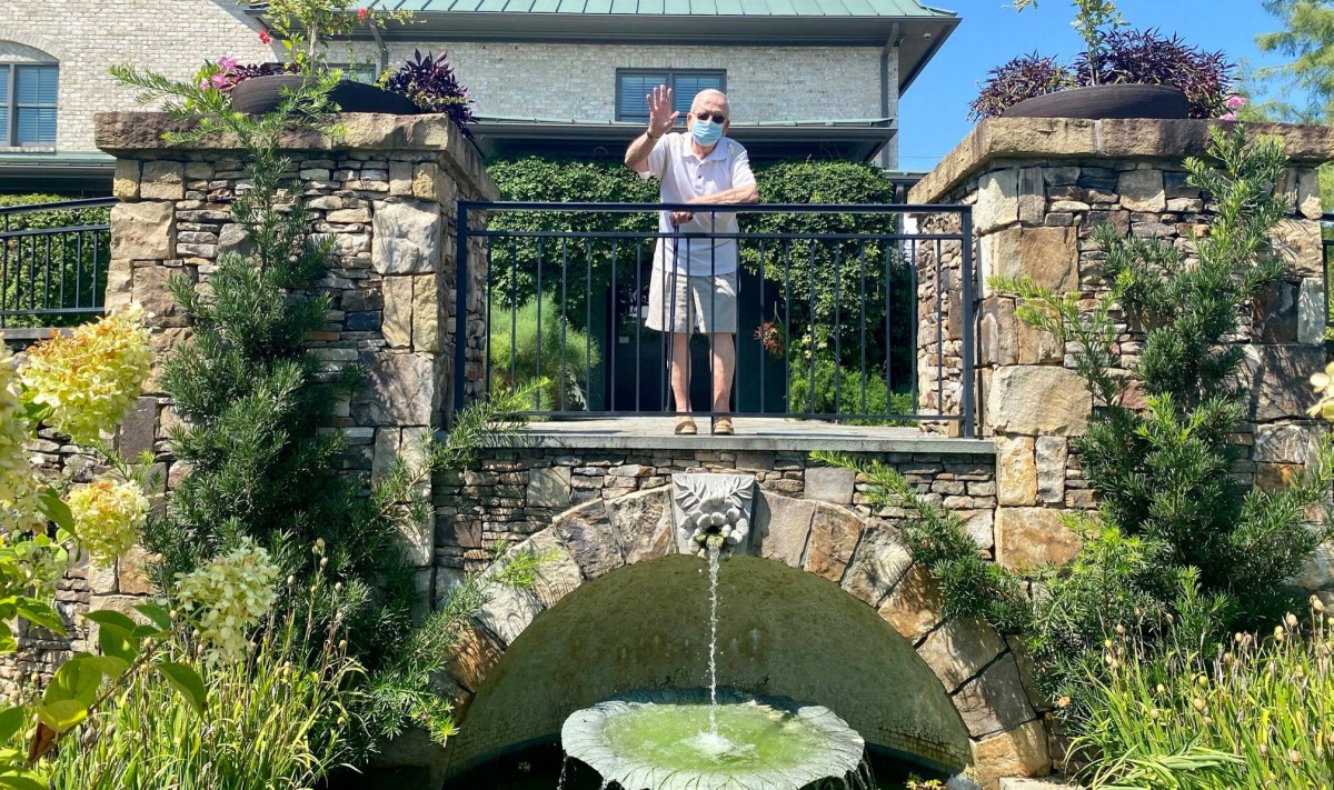 A person stands on a stone bridge above a fountain in a garden setting.