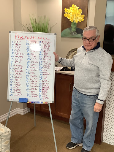 An elderly man proudly presents a list on a whiteboard, likely engaging in a community activity.