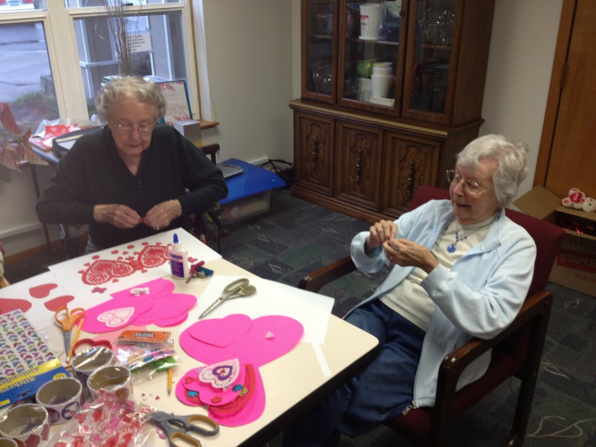 Two elderly individuals engaged in crafting activities at a table, surrounded by paper hearts and crafting materials.