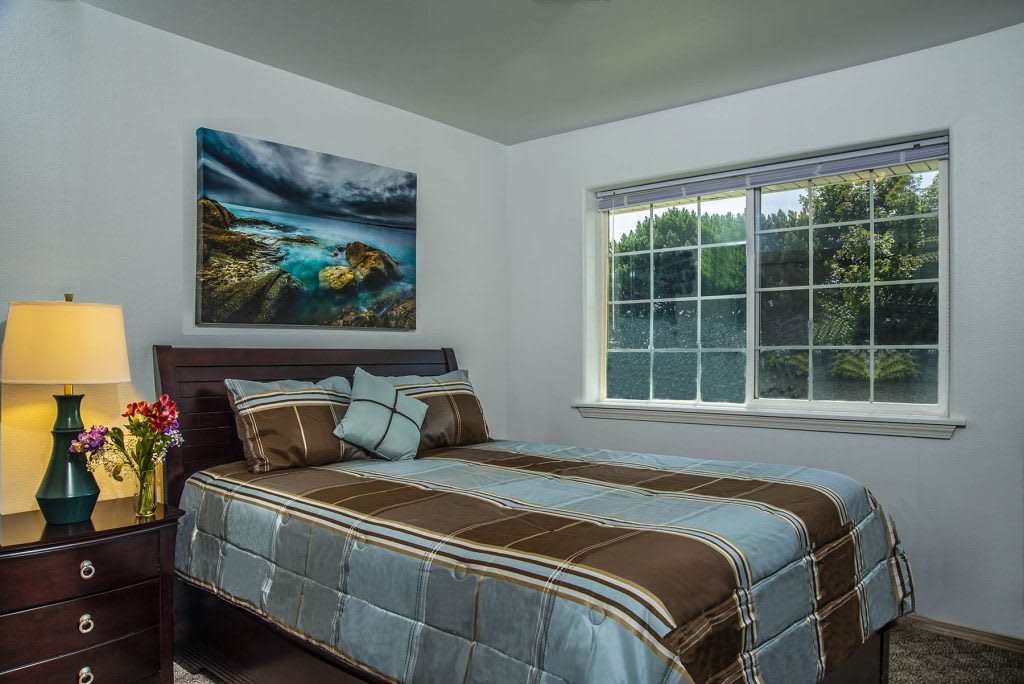 A cozy bedroom with a neatly made bed, a large window, and landscape artwork on the wall.