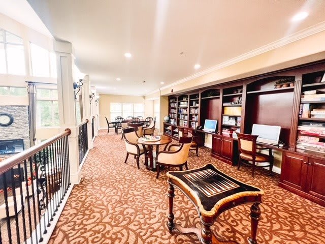 A well-lit library interior featuring computers, bookshelves, tables, and chairs arranged on a patterned carpet floor.