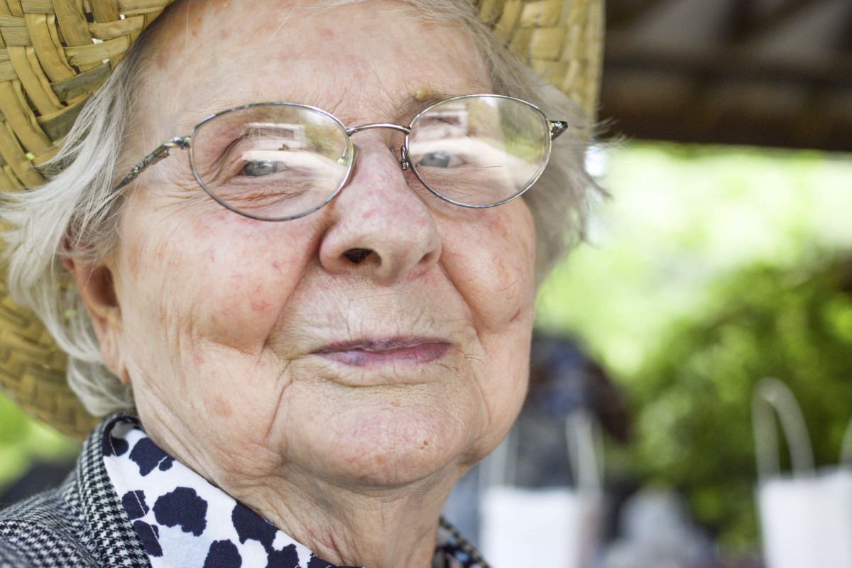 An elderly woman wearing glasses and a hat smiles warmly outdoors.