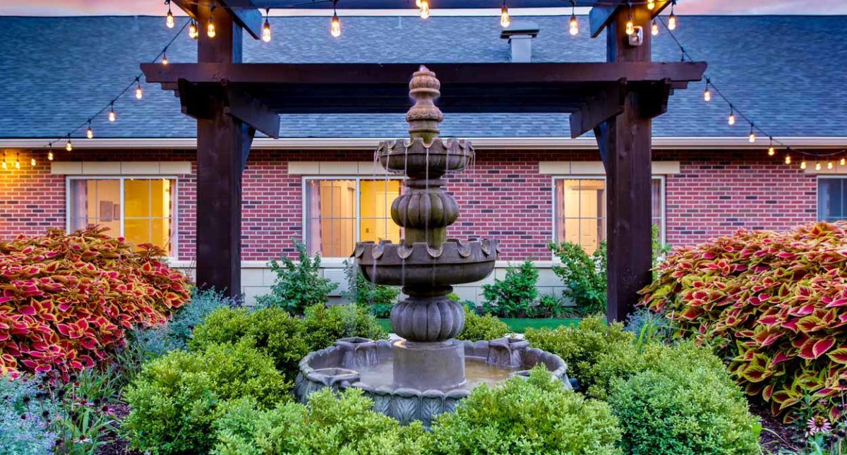 A decorative tiered fountain surrounded by lush greenery in front of a brick building, illuminated by string lights.