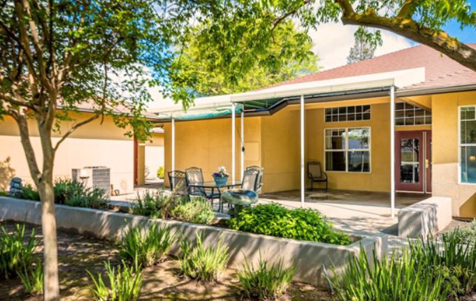 A cozy patio area with outdoor furniture surrounded by a garden and shaded by trees.
