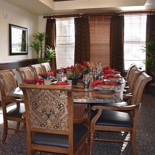 An elegantly set dining room with a large table and ornate chairs, featuring decorative plants and windows in the background.