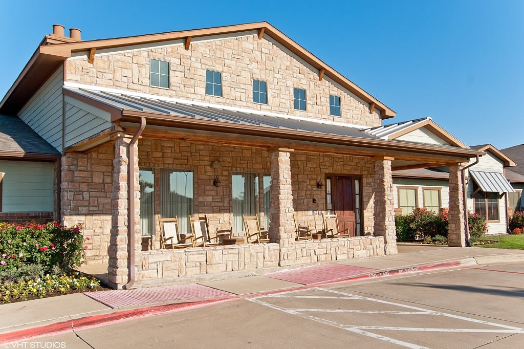 A brick building with a porch and rocking chairs, adjacent to a parking lot on a sunny day.