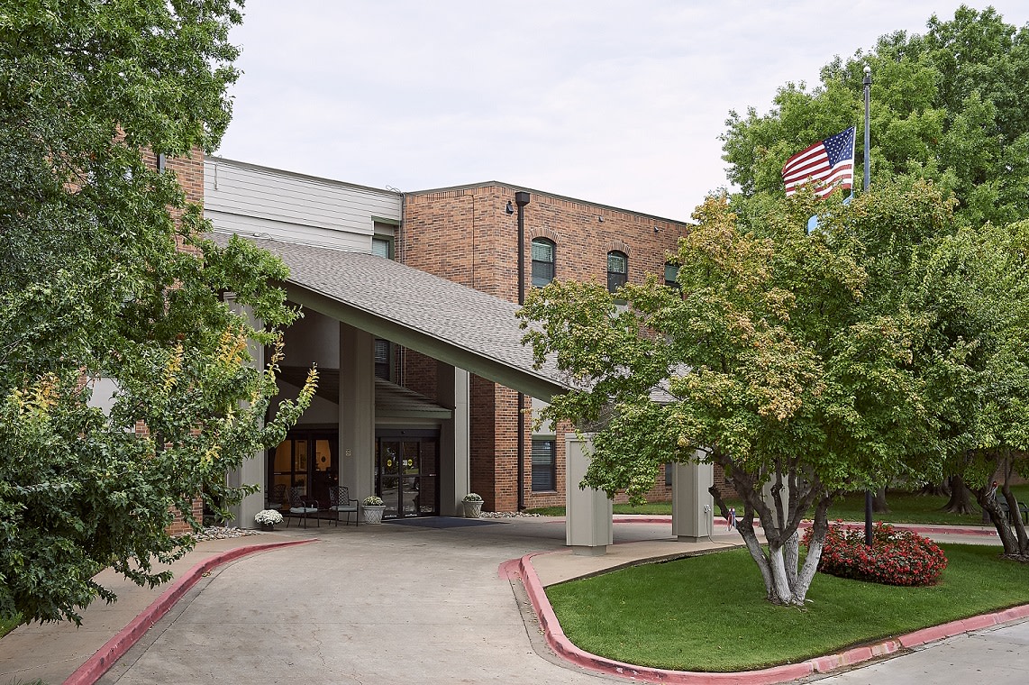 A view of a brick building entrance surrounded by trees, with an American flag flying nearby.