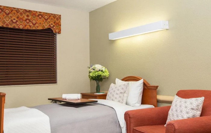 A well-lit hospital room with a neatly made bed and flowers on the bedside table.