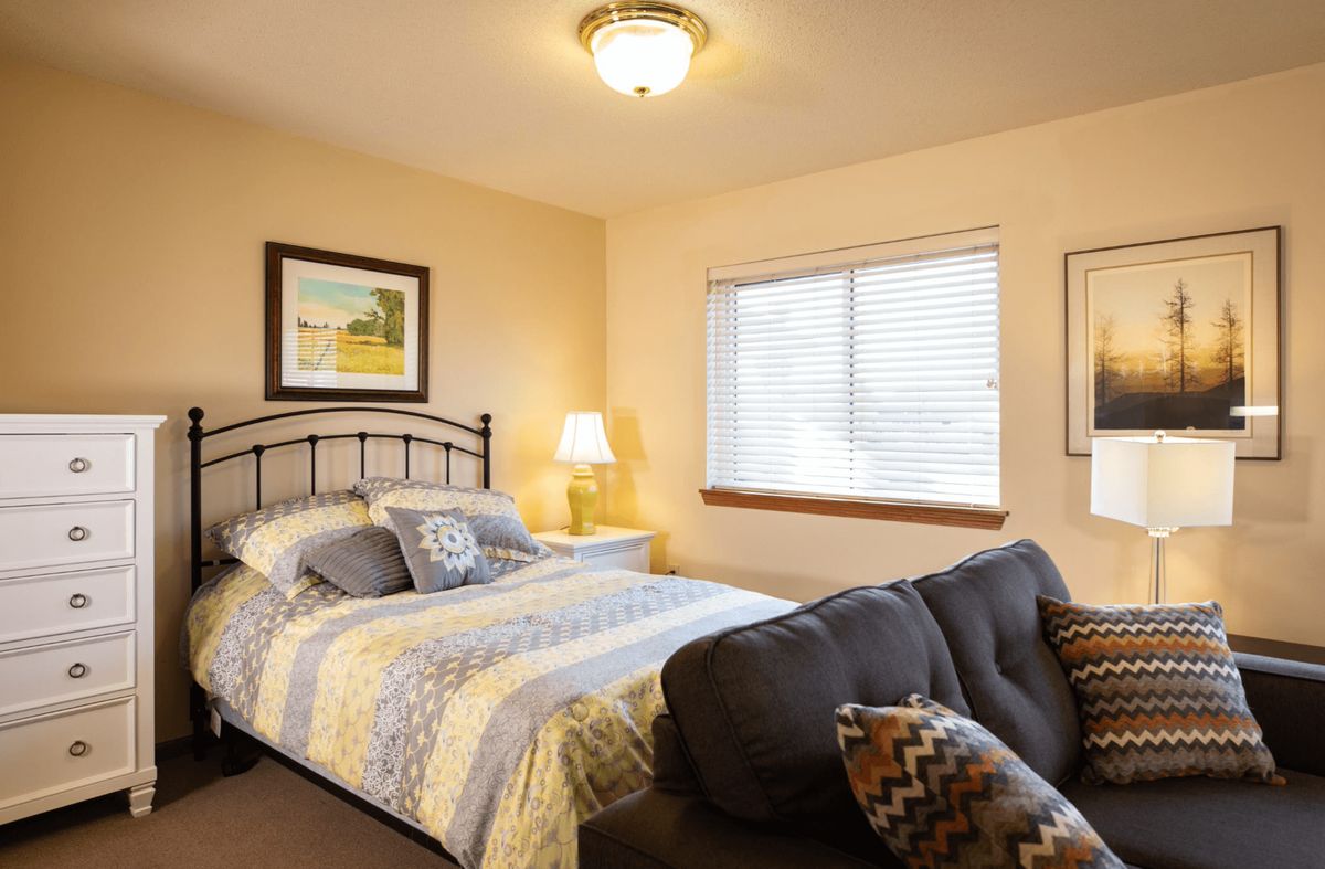 A cozy bedroom with a neatly made bed, a nightstand lamp, and a window allowing natural light in.