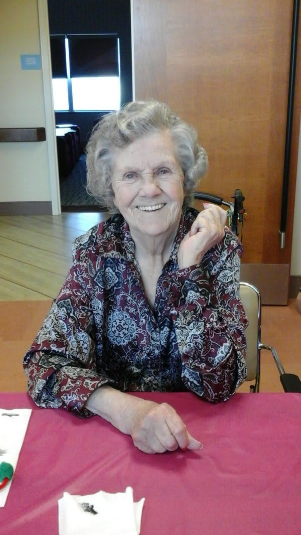 An elderly woman smiling while seated indoors, wearing a floral shirt.