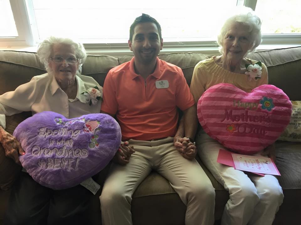 A group of three individuals sitting on a sofa, with the two elderly women holding heart-shaped pillows.