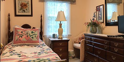 A cozy bedroom with floral bedding, a wooden dresser, and a lamp by the window.
