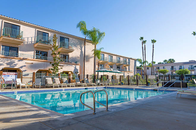 A tranquil outdoor swimming pool area in a hotel setting, surrounded by palm trees and lounge chairs on a sunny day.