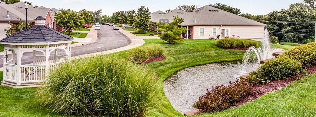 A peaceful suburban landscape featuring houses, a winding road, and a small pond surrounded by lush greenery.