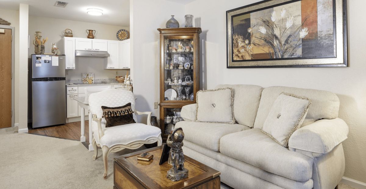 A cozy living room with a beige sofa, armchair, and a wooden coffee table adorned with decorative items. A glass cabinet displays collectibles, while artwork hangs on the wall.