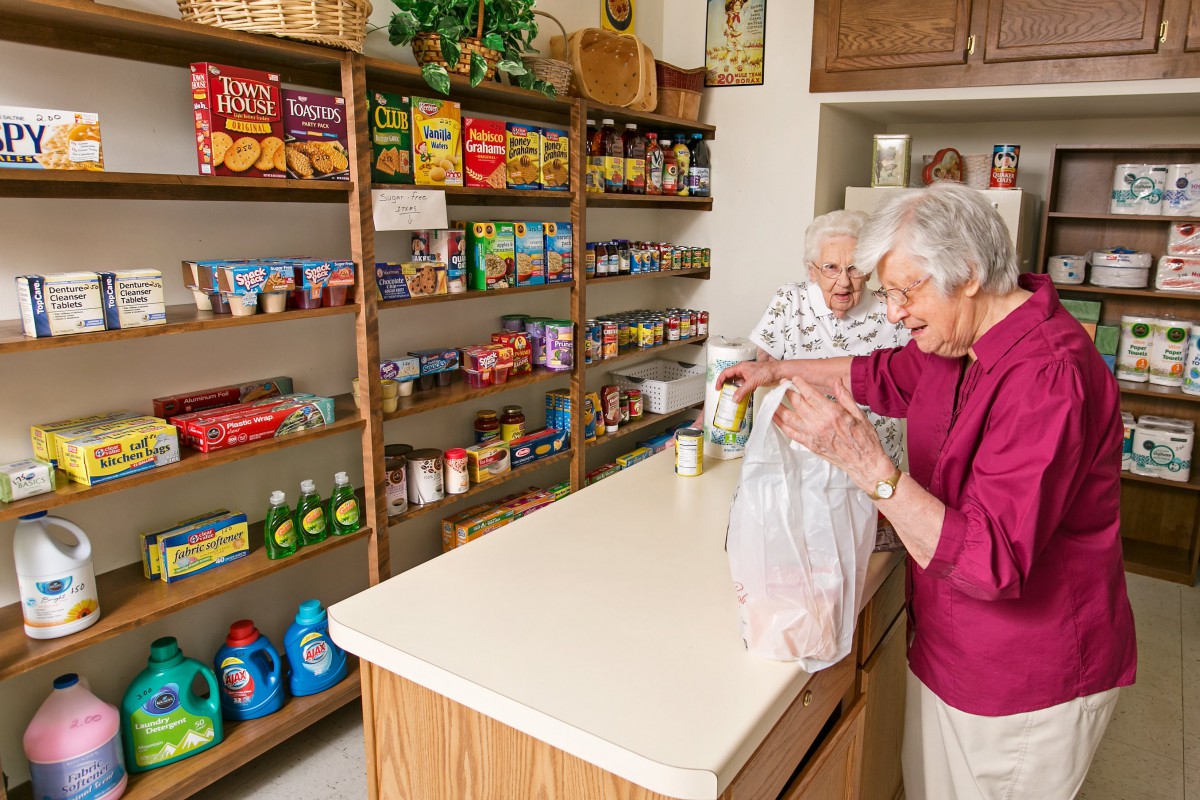 Two elderly women shopping and packing groceries at a small store, surrounded by shelves stocked with various food items.