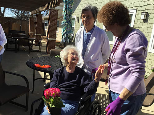 An elderly person in a wheelchair is outdoors, accompanied by two caregivers. They are enjoying a sunny day and holding a pot of flowers.