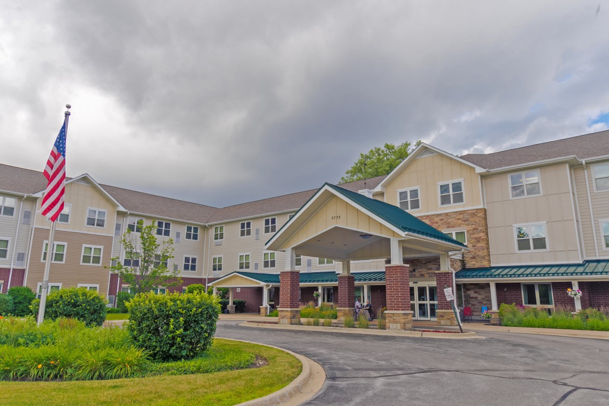 A large residential building with a covered entrance and surrounding greenery under a cloudy sky.
