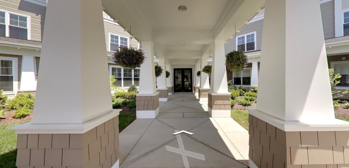 A covered walkway leads to the entrance of a building, flanked by decorative columns and landscaping.