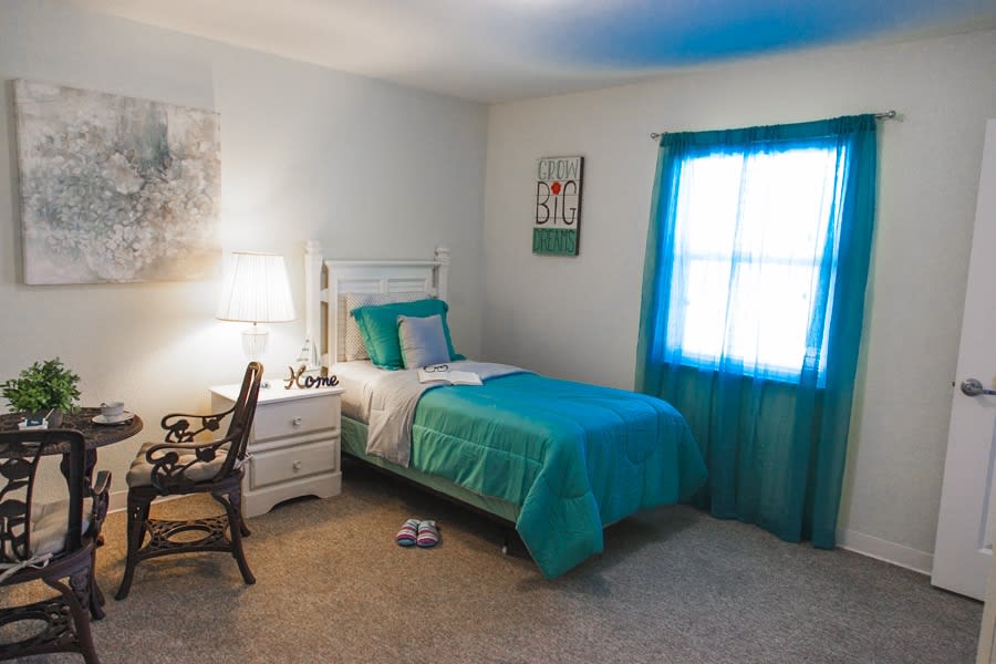 A cozy bedroom with a single bed dressed in turquoise bedding, featuring a nightstand, a lamp, and a window with matching curtains.